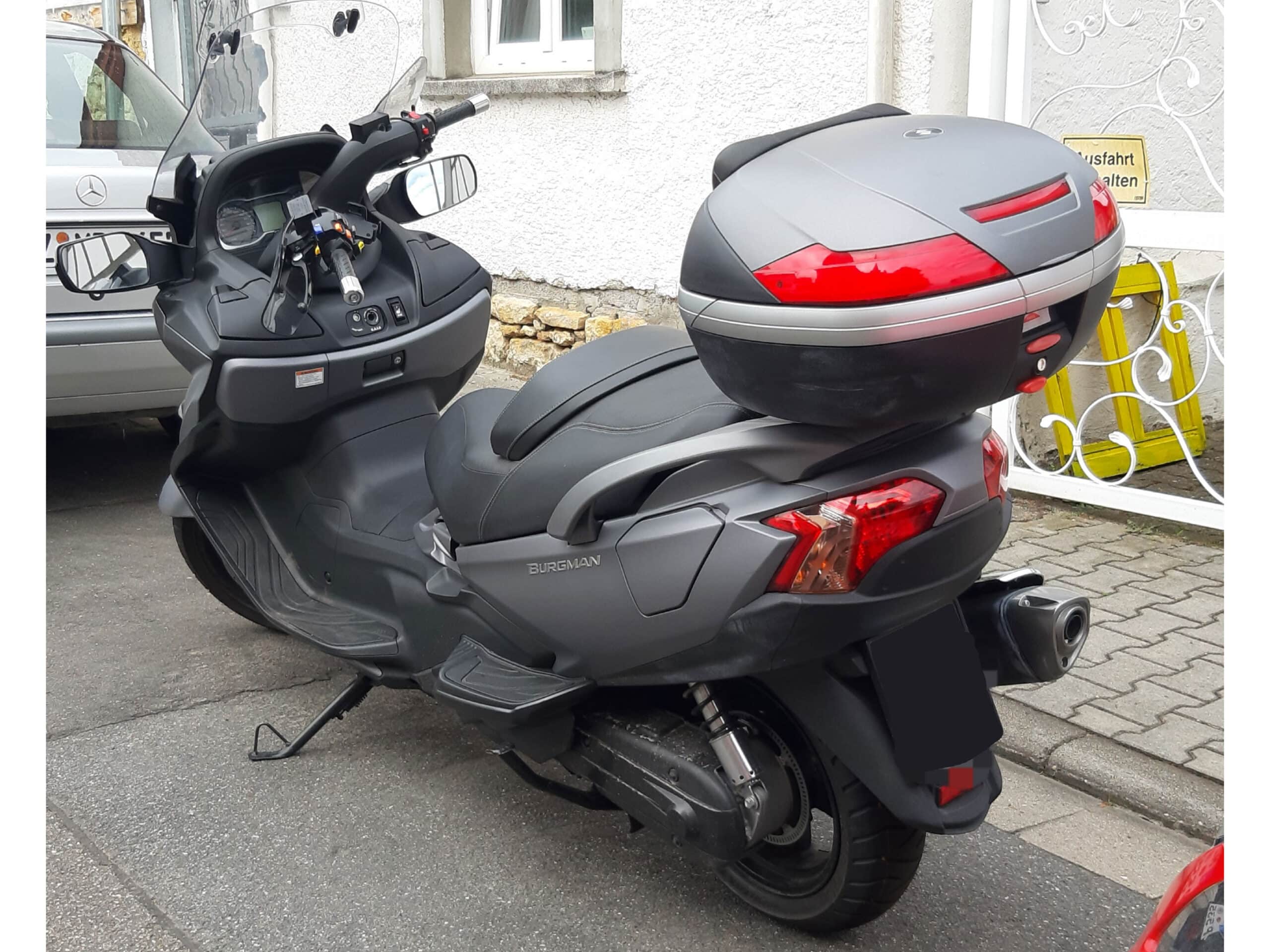 Gray Suzuki AN 650 BURGMAN ABS (2018) with top case on center stand, Scooter parked in front of a residential building.
